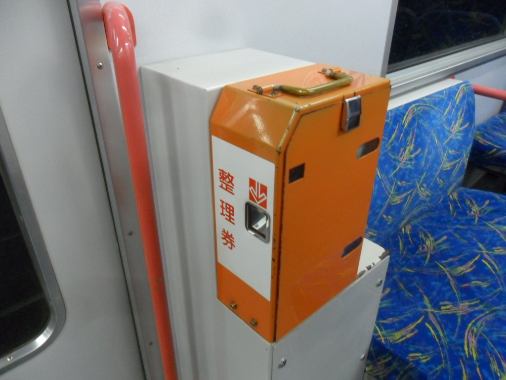 Close-up of an orange 整理券 (seiri-ken) numbered-ticket dispenser mounted at the doorway of a Japanese Meitetsu rail car